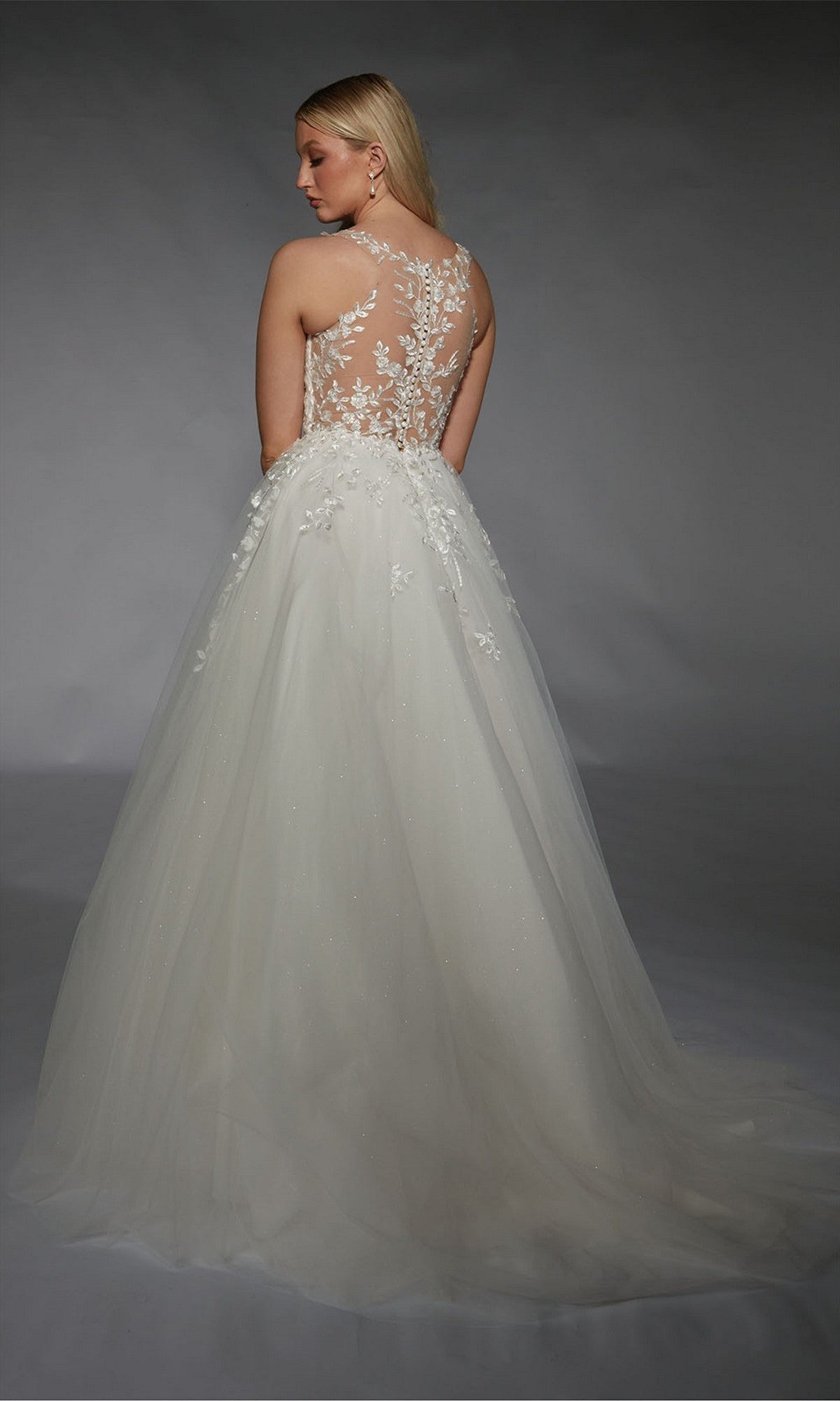 A woman stands facing away against a gray background, wearing the Long White Dress: Alyce 7128, a bridal gown featuring a sheer floral lace appliqué back and a long tulle skirt.